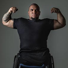 Foto de homem negro cadeirante, careca, vestido com camisa e bermuda preta, mostrando os músculos dos braços.